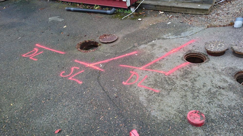 Asfalterad yta med två öppnade brunnslock och rosa sprejmarkeringar som visar avloppets sträckning.
